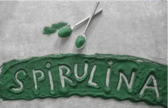 the word "Spirulina" on a green textured surface with two spoons filled with green spirulina powder above it